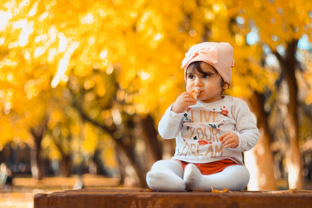 A cute baby girl sitting on a bench enjoying a sunny fall day in a park.