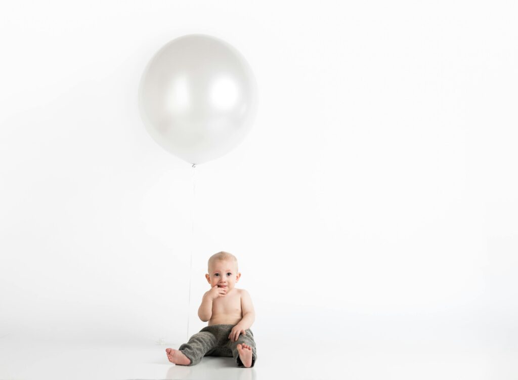 Cute baby sitting indoors with a large balloon in a minimalist white background.