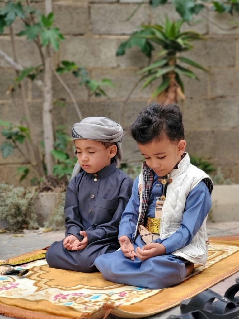 Two young boys in traditional attire praying outdoors in Sana'a, Yemen, showcasing cultural devotion.