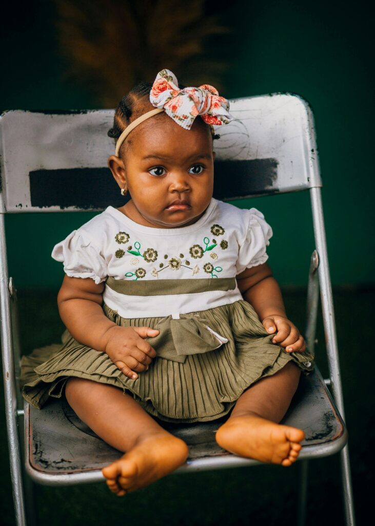 Cute African baby girl sitting on a chair outdoors, wearing a floral dress and bow.