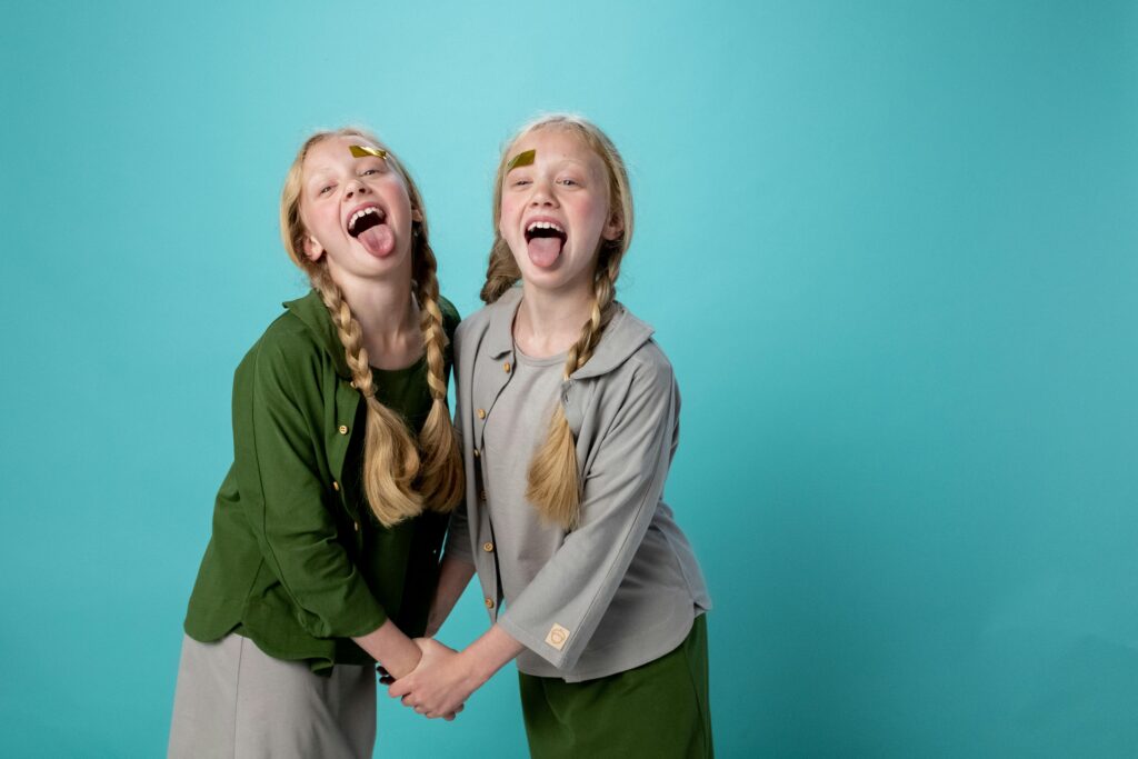 Two young twin girls with braids and face stickers posing playfully indoors against a turquoise background.