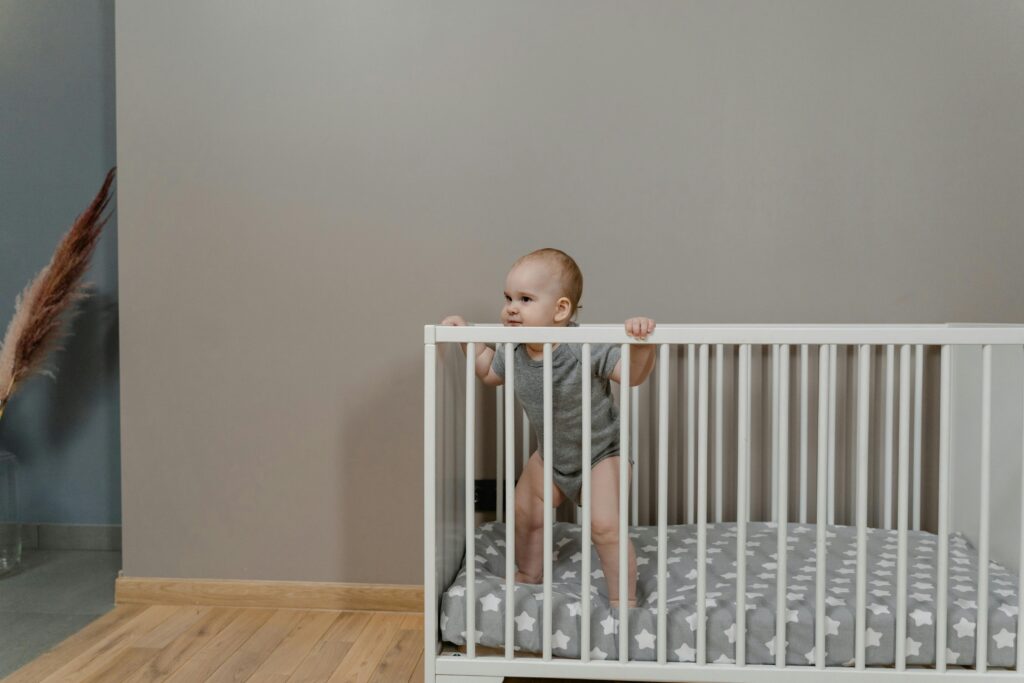 A cute baby in a grey onesie stands in a white crib indoors.