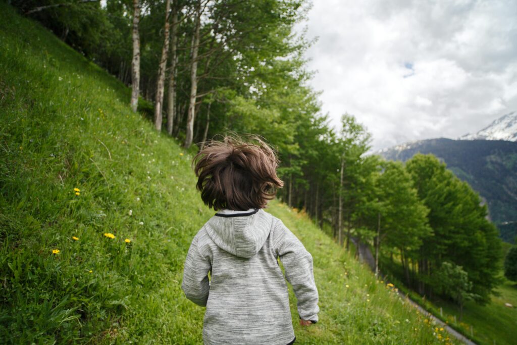 A young boy wearing a hoodie runs through a lush, mountainous landscape filled with trees and wildflowers.