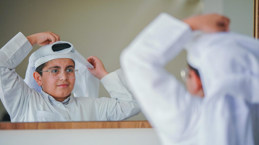 Middle Eastern boy adjusting his traditional attire in front of a mirror, indoors.